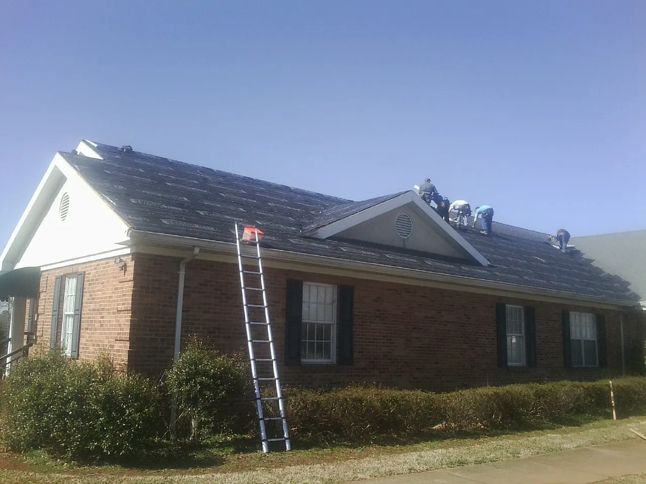 Crew tearing shingles off a brick ranch roof with a ladder set — Emergency Roof Repair work by our crew in La Junta
