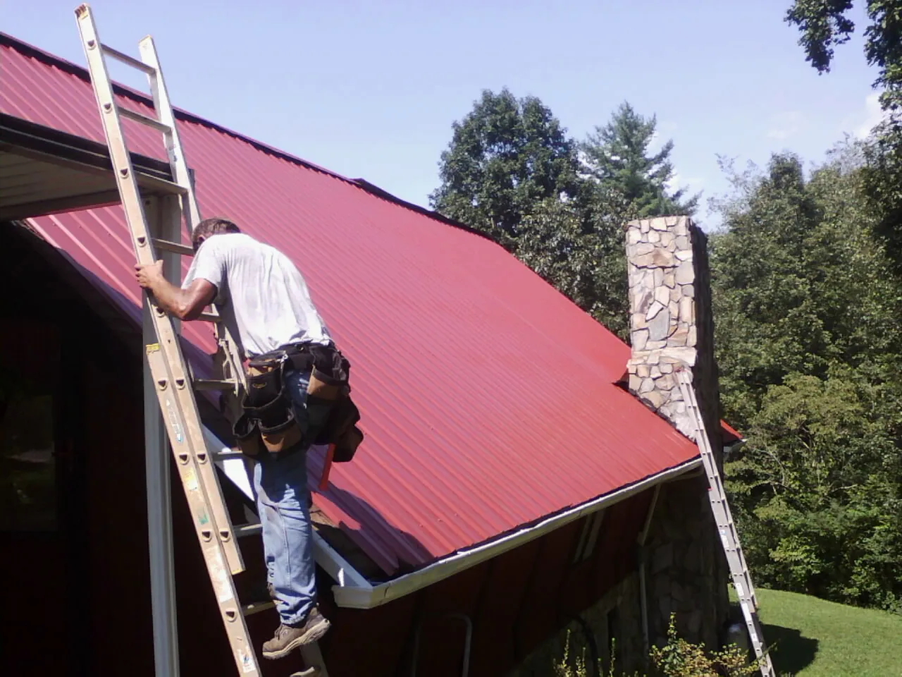 Roofer on a ladder beside a red metal roof and stone chimney — Soffit & Fascia Repair by Roofing Brothers in La Junta